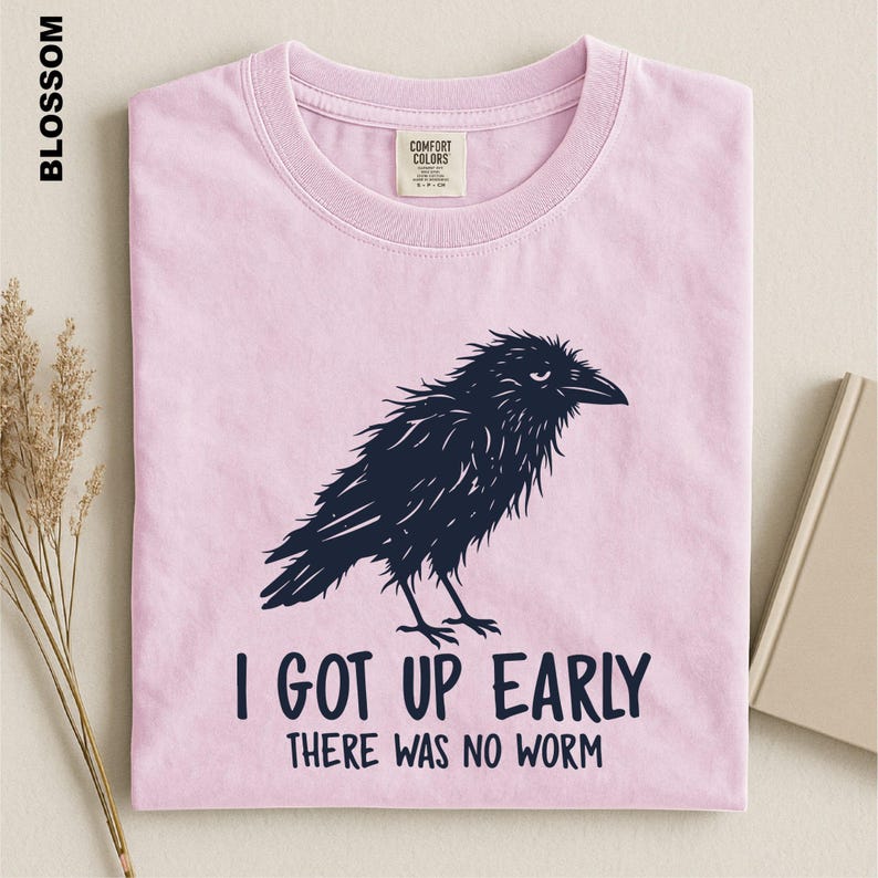 May include: A light pink short-sleeve t-shirt featuring a dark navy blue graphic of a ruffled crow. Below the bird, the text reads: I GOT UP EARLY THERE WAS NO WORM. The shirt has a crew neckline and a Comfort Colors brand label at the inner neck.