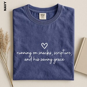 May include: A navy blue short-sleeve t-shirt with a crew neckline. The center of the shirt features a white heart icon above the white text: running on snacks, scripture, and his saving grace. The shirt has a Comfort Colors brand label.
