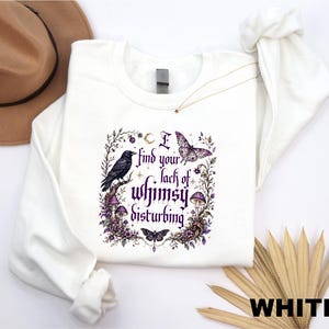 Puede incluir: Una sudadera blanca de manga larga con un estampado gr&aacute;fico que muestra un cuervo negro, una polilla, una mariposa y setas moradas. El texto dice I find your lack of whimsy disturbing, rodeado de elementos florales y celestiales en morado.