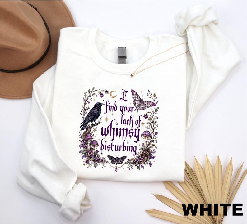Puede incluir: Una sudadera blanca de manga larga con un estampado gr&aacute;fico que muestra un cuervo negro, una polilla, una mariposa y setas moradas. El texto dice I find your lack of whimsy disturbing, rodeado de elementos florales y celestiales en morado.