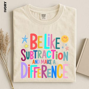 May include: An ivory-colored crew neck t-shirt featuring the multi-colored text "BE LIKE SUBTRACTION AND MAKE A DIFFERENCE" printed on the front. The design includes various small star shapes and a yellow smiley face icon in vibrant colors.