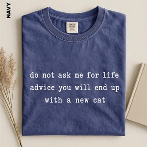 May include: A navy blue short-sleeved t-shirt with a crew neckline. The shirt features white text printed on the front that reads: do not ask me for life advice you will end up with a new cat. The garment has a Comfort Colors brand label.
