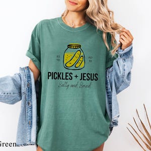 A woman wearing a green t-shirt with a graphic of a jar of pickles and the text "PICKLES + JESUS Salty and Saved" printed on it.