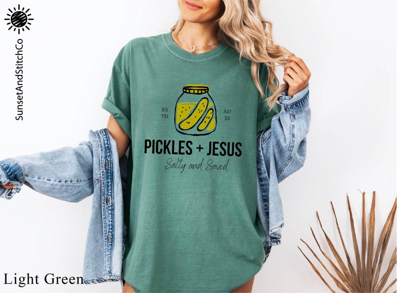 A woman wearing a green t-shirt with a graphic of a jar of pickles and the text "PICKLES + JESUS Salty and Saved" printed on it.