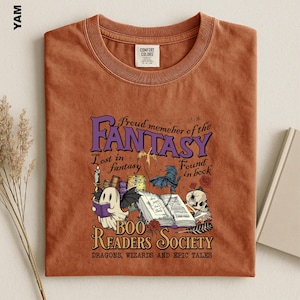 May include: A rust-colored crew neck t-shirt featuring a graphic print with the text Proud memeher of the FANTASY, Lost in fantasy, Found in book, BOO READERS SOCIETY, DRAGONS, WIZARDS AND EPIC TALES, a ghost, a dragon, a skull, and open books.
