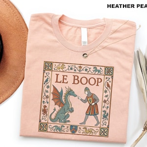 May include: A heather peach t-shirt featuring a medieval-style graphic of a knight touching a dragon's nose with the text LE BOOP. The design is enclosed in a decorative floral border with small mythical creatures and birds in muted tones.