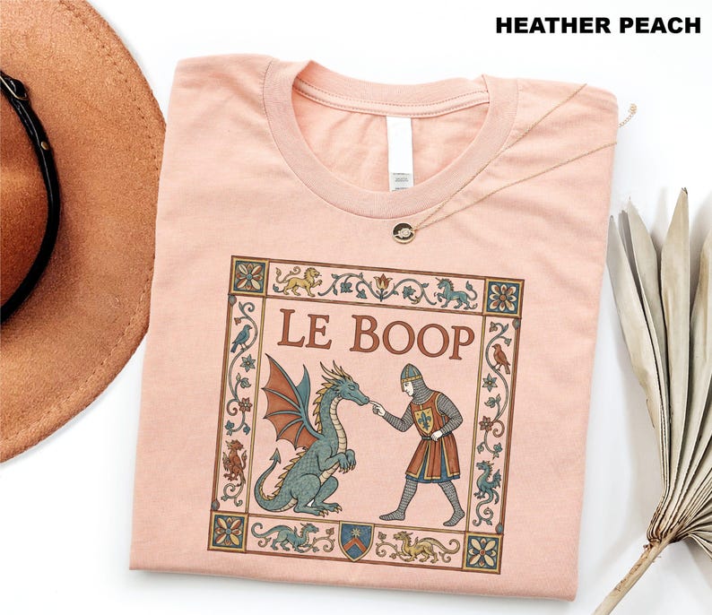May include: A heather peach t-shirt featuring a medieval-style graphic of a knight touching a dragon's nose with the text LE BOOP. The design is enclosed in a decorative floral border with small mythical creatures and birds in muted tones.