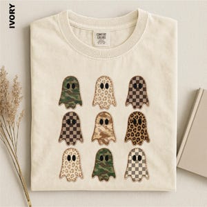 May include: An ivory short-sleeve t-shirt featuring a graphic print of nine ghosts arranged in a three-by-three grid. Each ghost displays a different pattern, including camouflage, leopard print, and brown checkerboard. The shirt has a crew neckline.