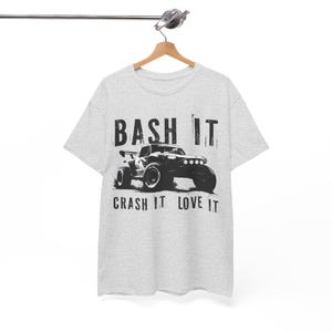 Peut inclure: T-shirt gris clair avec un dessin noir d'un monster truck et le texte "BASH IT" au-dessus et "CRASH IT LOVE IT" en dessous. Le t-shirt est suspendu à un cintre en bois.