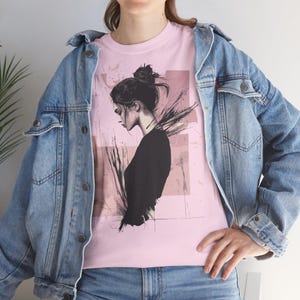 Op de afbeelding: Lichtroze T-shirt met een zwart-witte illustratie van een vrouw met haar haar in een knot. De afbeelding heeft een moderne, artistieke stijl met abstracte penseelstreken op de achtergrond. De persoon draagt een lichtblauwe spijkerjas.