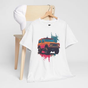 Op de afbeelding: Wit t-shirt met een oranje en zwarte terreinwagenafbeelding en kleurrijke verfspat-accenten. Het shirt hangt aan een houten hanger. Een witte baseballcap rust op een houten stoel.