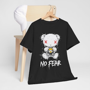 Op de afbeelding: Zwart t-shirt met een witte afbeelding van een teddybeer met rode 'X'-ogen, die een geel stuur vasthoudt. De woorden 'NO FEAR' staan eronder in het wit gedrukt. Een witte baseballcap hangt aan een houten stoel.