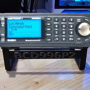 Puede incluir: Un escáner de radio Uniden Bearcat TrunkTracker V negro con una pantalla LCD azul que muestra "Los Almos Command Post" y un soporte negro con el texto "BCD996P2" impreso en él.