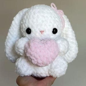Puede incluir: Un conejo de peluche tejido a ganchillo en color blanco con orejas largas, ojos de plástico negro y una pequeña nariz bordada en rosa. El conejo sostiene un corazón tejido en rosa claro entre sus patas delanteras y un lazo rosa en la cabeza.