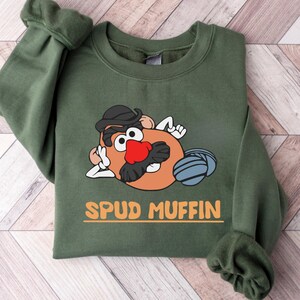 Puede incluir: Sudadera verde con un dibujo de un personaje de dibujos animados de patata con sombrero y bigote, con el texto "SPUD MUFFIN" en naranja. La sudadera está doblada sobre una superficie de madera clara.