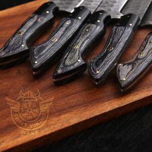 May include: A set of kitchen knives with dark wood grain handles and silver blades. The handles are accented with gold-colored rivets and a decorative emblem. The knives are arranged on a wooden cutting board.