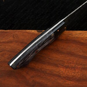 May include: Close-up of a knife with a dark, textured wooden handle and a silver blade. The handle has a black and gray wood grain pattern. The blade is silver with a sharp edge. The knife is on a wooden surface.