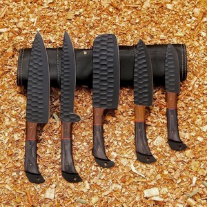 Puede incluir: Un juego de cinco cuchillos de chef negros con hojas texturizadas y mangos de madera. Los cuchillos se muestran en un estuche de cuero negro, sobre un fondo de virutas de madera. Las hojas tienen una textura martillada &uacute;nica.