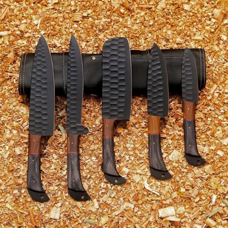 Puede incluir: Un juego de cinco cuchillos de chef negros con hojas texturizadas y mangos de madera. Los cuchillos se muestran en un estuche de cuero negro, sobre un fondo de virutas de madera. Las hojas tienen una textura martillada &uacute;nica.