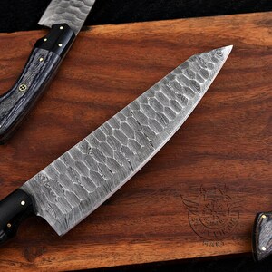 May include: Close-up of a chef's knife with a patterned blade and a black handle. The knife is on a wooden cutting board. The blade has a unique textured design. The handle is black with gold accents.