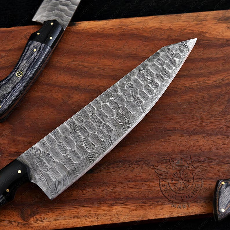 May include: Close-up of a chef's knife with a patterned blade and a black handle. The knife is on a wooden cutting board. The blade has a unique textured design. The handle is black with gold accents.