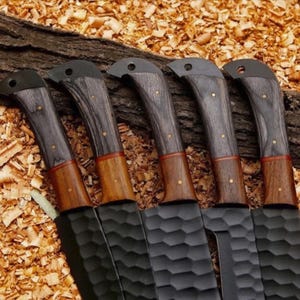 Puede incluir: Cinco cuchillos negros con mangos de madera. Los mangos son una combinaci&oacute;n de madera gris oscuro y marr&oacute;n, con remaches de lat&oacute;n. Las hojas tienen una superficie texturizada. Los cuchillos est&aacute;n dispuestos sobre un lecho de virutas de madera.