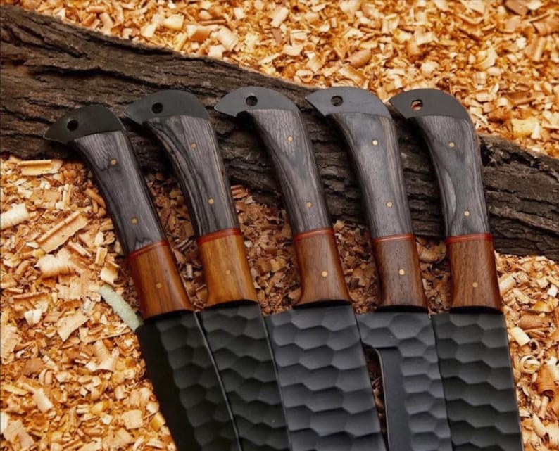 Puede incluir: Cinco cuchillos negros con mangos de madera. Los mangos son una combinaci&oacute;n de madera gris oscuro y marr&oacute;n, con remaches de lat&oacute;n. Las hojas tienen una superficie texturizada. Los cuchillos est&aacute;n dispuestos sobre un lecho de virutas de madera.