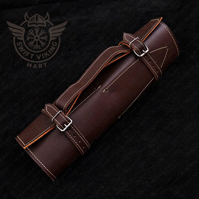May include: A rolled-up brown leather knife roll with white stitching and silver buckles. The roll has a handle and a strap for securing the contents. The Swift Viking Mart logo is visible in the upper left corner.