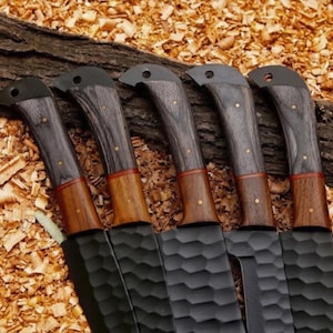 Puede incluir: Cinco cuchillos negros con mangos de madera. Los mangos son una combinaci&oacute;n de madera gris oscuro y marr&oacute;n, con remaches de lat&oacute;n. Las hojas tienen una superficie texturizada. Los cuchillos est&aacute;n dispuestos sobre un lecho de virutas de madera.