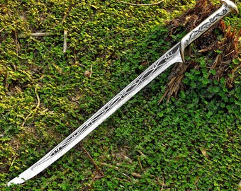 May include: A silver fantasy sword with intricate black etched designs on the blade and handle. The sword is lying on a bed of green foliage, showcasing its detailed craftsmanship and fantasy theme.