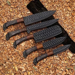 Puede incluir: Un juego de cinco cuchillos de cocina negros con hojas texturizadas y mangos de madera. Los mangos son de color marr&oacute;n oscuro con un acento rojo cerca de la hoja. Los cuchillos est&aacute;n colocados sobre un lecho de virutas de madera.