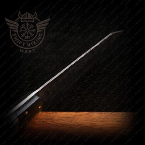 May include: A black-handled knife with a long, silver blade. The blade has a textured surface and is angled. The knife is set against a dark background with a wooden surface at the bottom. The Swift Viking Mart logo is in the top left corner.