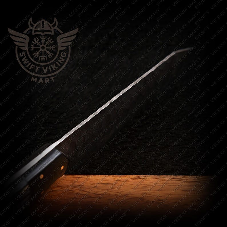 May include: A black-handled knife with a long, silver blade. The blade has a textured surface and is angled. The knife is set against a dark background with a wooden surface at the bottom. The Swift Viking Mart logo is in the top left corner.