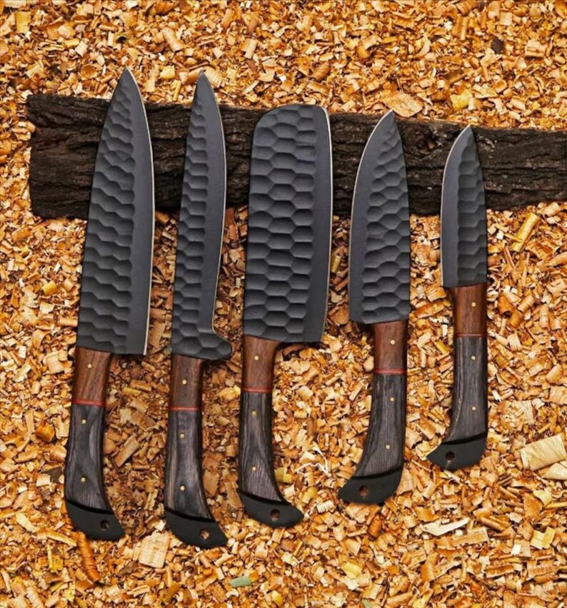 Puede incluir: Un juego de cinco cuchillos de cocina con hojas de color gris oscuro y mangos de madera. Las hojas tienen una superficie texturizada y los mangos combinan madera marr&oacute;n y negra. Los cuchillos est&aacute;n colocados sobre un lecho de virutas de madera.