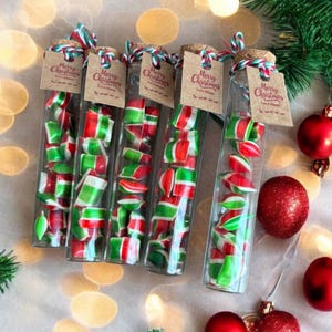 May include: Several glass tubes filled with red, green, and white striped candies. Each tube has a cork stopper and a small tag that reads "Merry Christmas". Red Christmas ornaments and greenery are arranged around the tubes, creating a festive holiday scene.