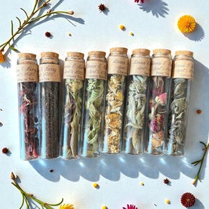May include: A collection of glass vials filled with various dried herbs and teas, each sealed with a cork stopper and labeled. The labels include names like "Hibiscus Tea" and "Rose Tea." The vials are arranged on a white surface, adorned with flowers.