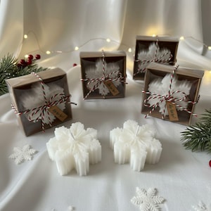 May include: White snowflake-shaped candles, some packaged in square brown boxes with clear windows, tied with red and white twine and small brown tags reading Ho ho ho!. Two loose snowflake candles sit in front of the boxed candles.