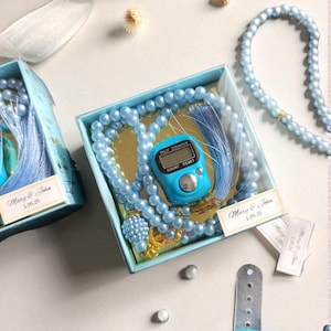 May include: A light blue prayer bead set with a digital tally counter, presented in a matching box. The set includes a beaded necklace and a counter displaying "135". The box is decorated with the text "Mary & John 1.05.25".