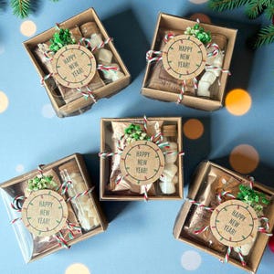 Könnte beinhalten: Fünf Geschenkboxen mit transparenten Deckeln, jede gefüllt mit Leckereien und einer kleinen Flasche. Jede Box ist mit einem runden Etikett mit der Aufschrift "Happy New Year!" verziert und mit rot-weiß-grünem Garn gebunden. Auf blauem Hintergrund.