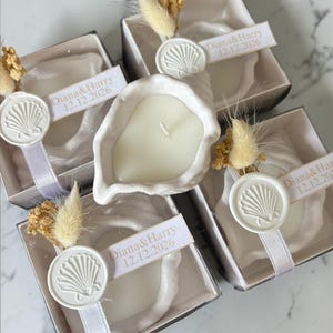 Può includere: Cinque candele a forma di conchiglia in scatole individuali. Ogni candela bianca è posta in una conchiglia di ceramica bianca. Le scatole sono decorate con un nastro bianco, un sigillo di cera a forma di conchiglia e un'etichetta con la scritta "Diana & Harry 12.12.2026".