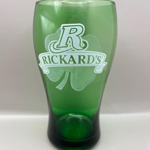 May include: Green glass with a shamrock design and the text "RICKARD'S". The glass has a rounded base and a wider top. The shamrock and text are in white. The glass is a vibrant green colour.