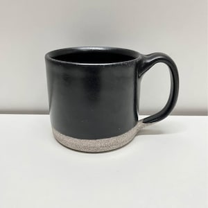 Può includere: Una tazza in ceramica caratterizzata da una finitura nera opaca e liscia sulla parte superiore e da una base testurizzata grigio chiaro maculata. La tazza presenta un manico arrotondato sul lato destro e un'apertura ampia, che evidenzia il netto contrasto tra lo smalto scuro e la sezione inferiore grezza e granulosa.