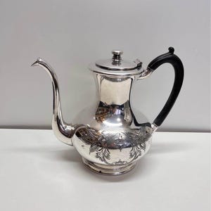 Peut inclure: Une théière argentée avec un long bec incurvé et une poignée noire. La théière a un couvercle et des gravures florales décoratives autour de la base. La théière est probablement en métal et est conçue pour servir des boissons.