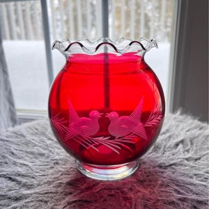 May include: A bright red glass candle holder with a wavy rim. The round body has an etched design of two birds facing each other. The candle holder sits on a clear, circular base, ideal for adding a touch of elegance to any room.