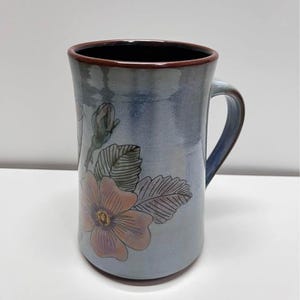 Puede incluir: Una taza de cerámica con un esmalte azul y un borde marrón. La taza presenta un diseño floral con una flor rosa y hojas verdes. El asa es azul y la taza es adecuada para bebidas frías o calientes.
