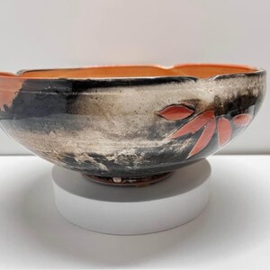 May include: A handmade ceramic bowl with a wide, irregular rim. The exterior features a blend of black, gray, and white tones, with accents of orange and red leaf designs. The interior is a warm orange color. This decorative bowl is suitable for serving or display.