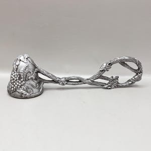 May include: Silver-colored serving tongs with a grape and vine design. The tongs feature a scoop-shaped end with a cluster of grapes and leaves, connected to a handle shaped like intertwined vines. The handle forms a loop at the end.