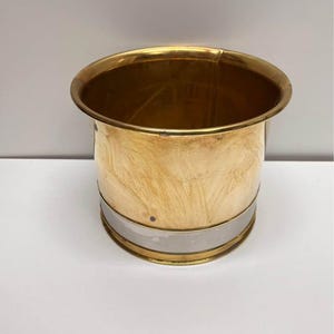 May include: A cylindrical brass container with a silver band around the middle. The container has a wide, rounded rim and a brushed finish. The interior is visible and appears to be empty. The container is sitting on a white surface.