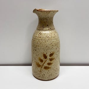 Puede incluir: Un jarrón de cerámica beige moteado con un diseño de hojas. El jarrón tiene un cuello estrecho y una parte superior ensanchada con un pequeño pico. El cuerpo del jarrón es cilíndrico y el diseño de hojas es de color marrón oscuro.