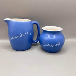 Może przedstawiać: Niebieski ceramiczny zestaw dzbanek i mały słoik. Dzbanek ma uchwyt i napis "Downderry". Słoik ma napis "Liskeard". Oba elementy mają białe wnętrze.